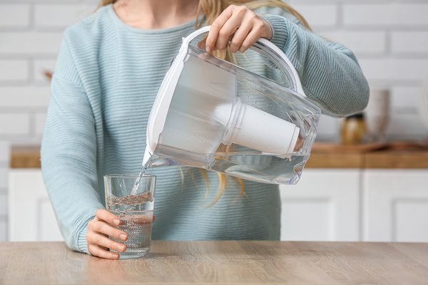 Woman pouring water into glass from filter jug at table in kitchen